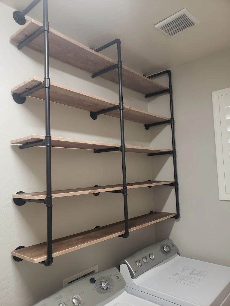 Industrial pipe shelving with five wooden shelves mounted above laundry appliances