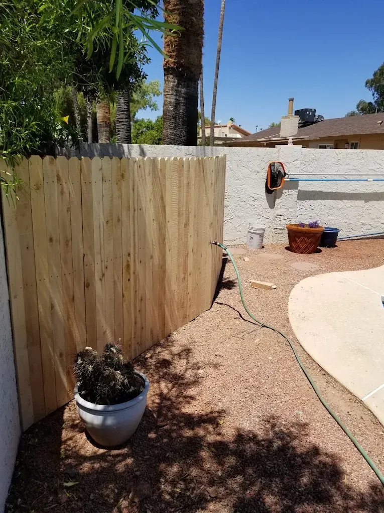 Residential backyard with newly installed wooden fence, concrete patio, potted cactus, and garden supplies under clear blue sky