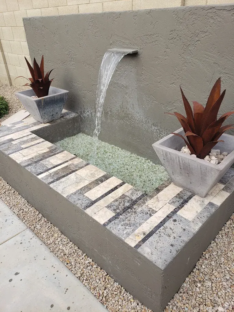 Modern water feature with cascading waterfall, stepping stones, green gravel, and red potted plants against concrete wall