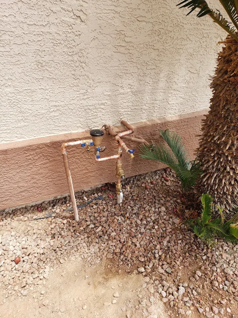 Copper water pipes with blue valves mounted on terracotta wall beside palm tree and gravel landscaping